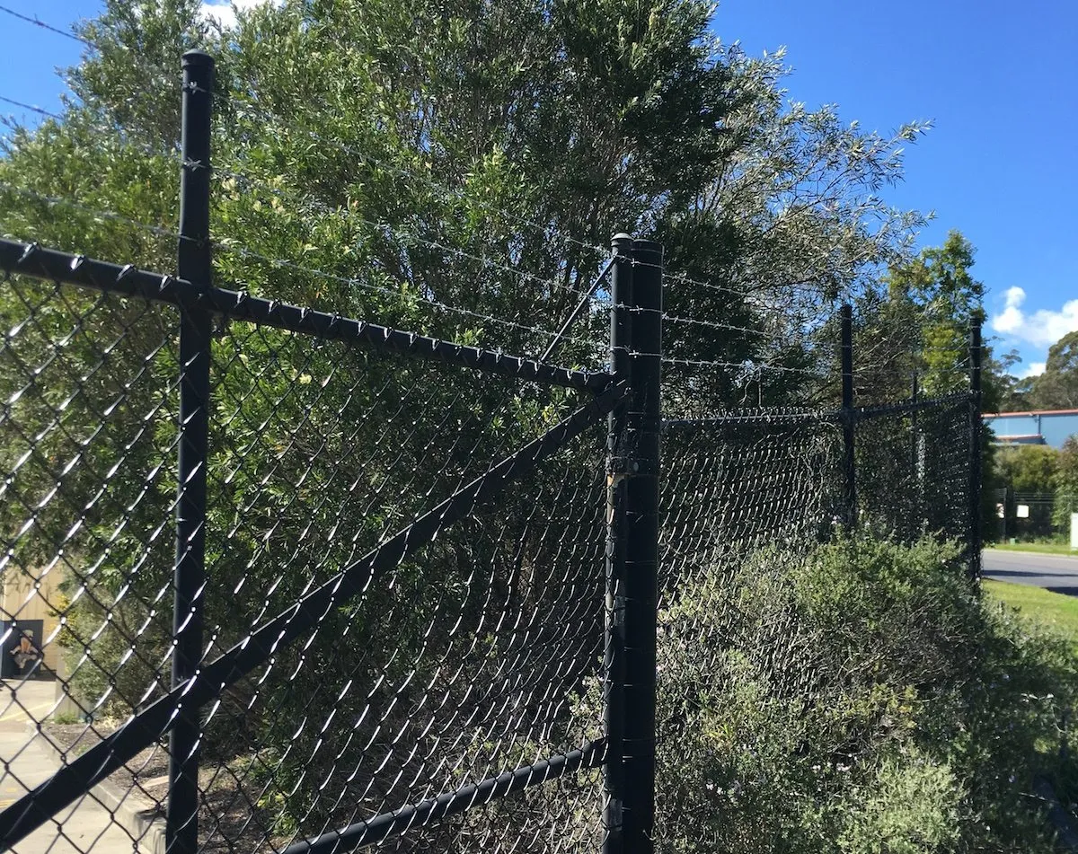 Black chainwire fence with a barbed-wire top along a roadside boundary