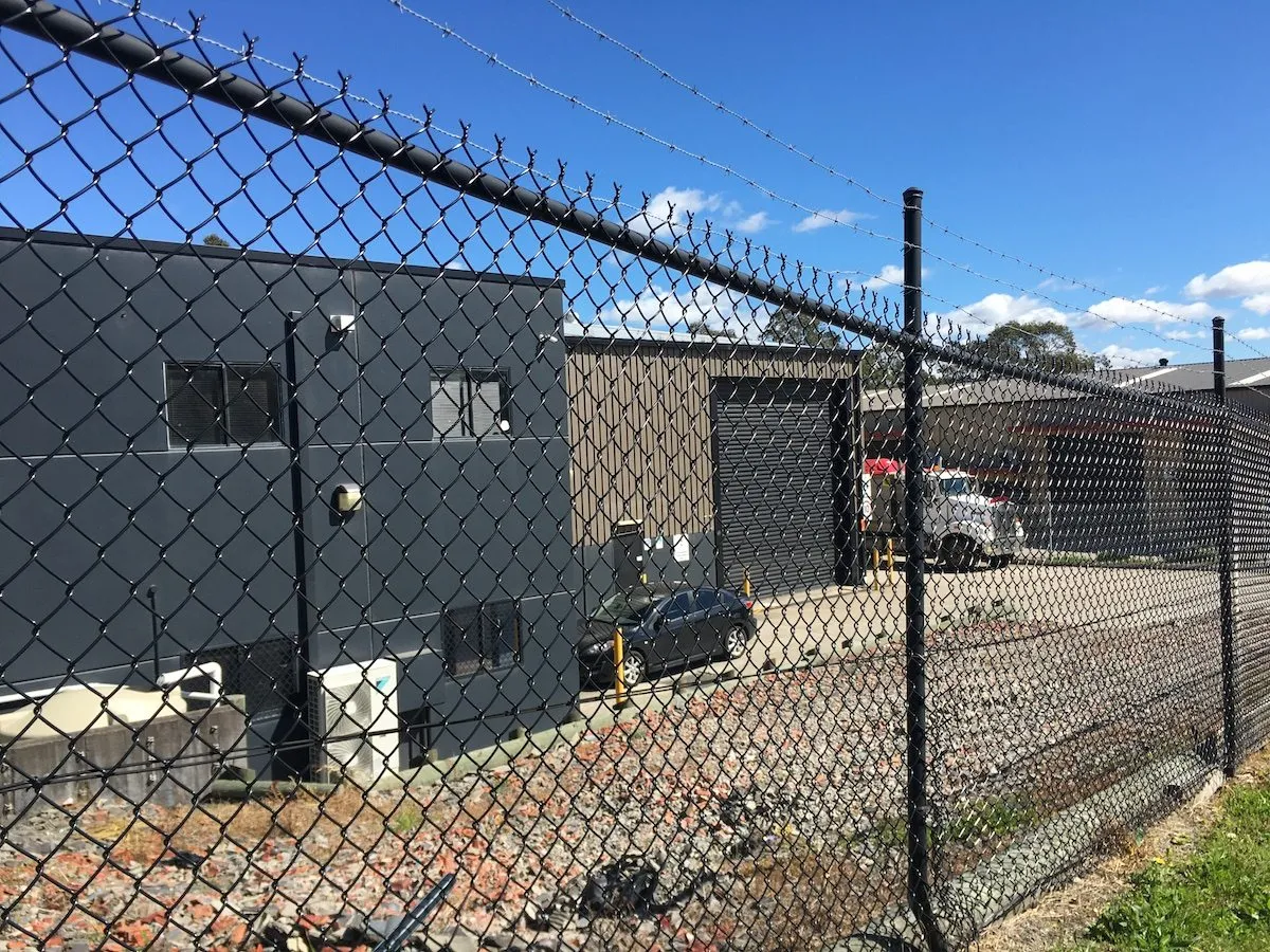 Black chainwire fence with a barbed-wire top around a commercial building