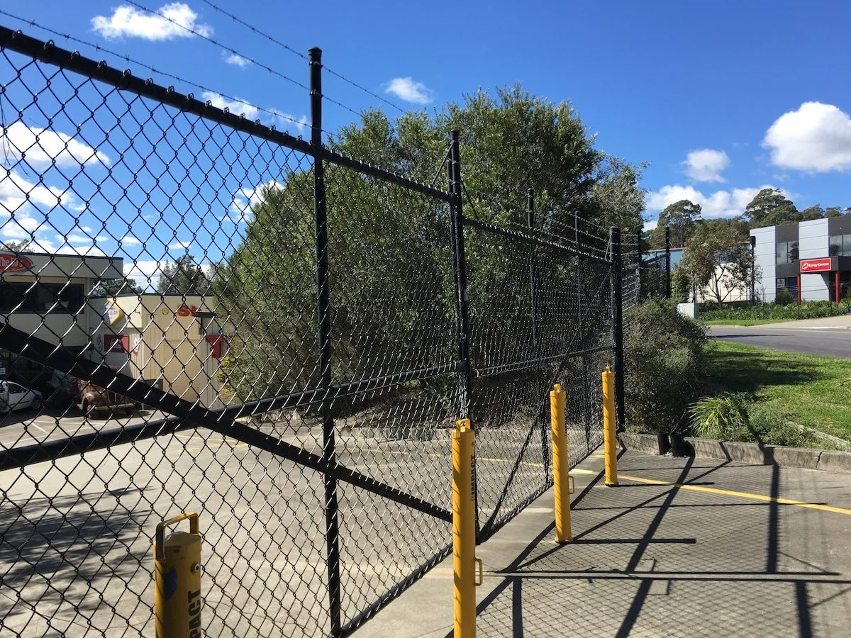 Black chainwire security fence with a barbed-wire top and bollards at a commercial site