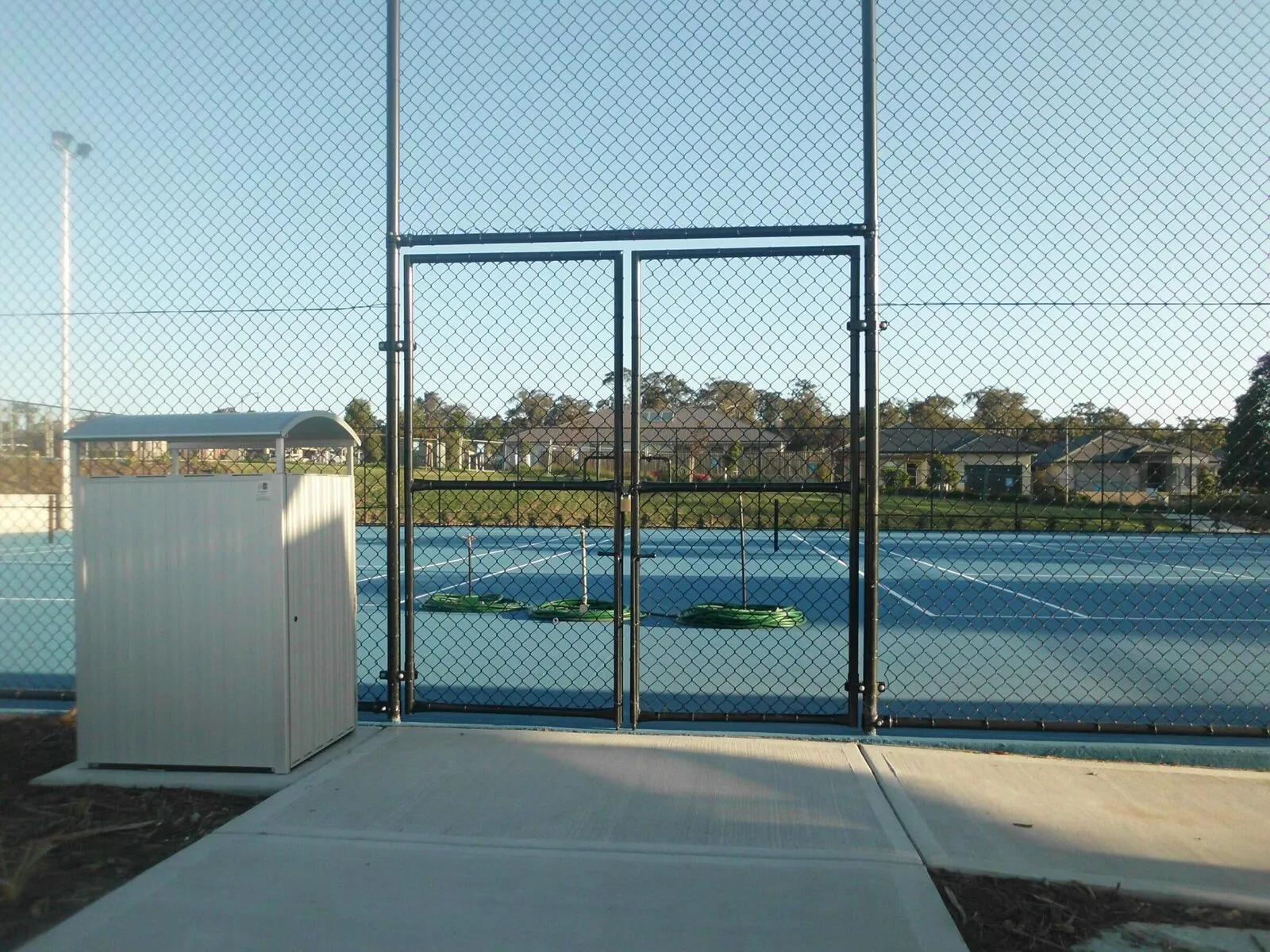 A high black chainwire fence enclosing a blue outdoor tennis court.