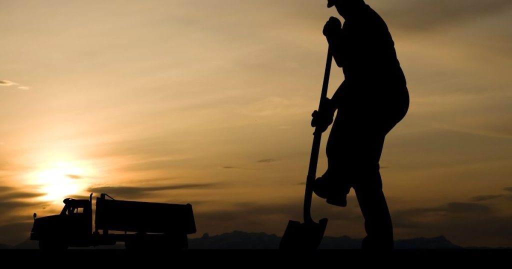 A silhouette of a construction worker using a shovel next to a dump truck against a sunset sky.