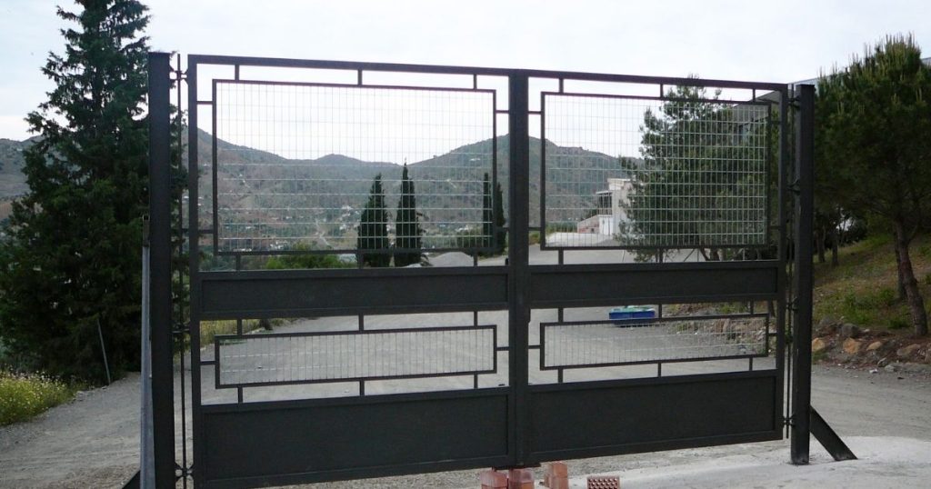 A large black metal gate with rectangular wire mesh inserts overlooking a hilly landscape and a dirt road.