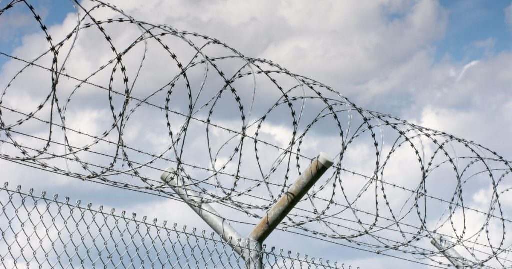 Coils of razor wire mounted on top of a chain-link fence against a cloudy sky.