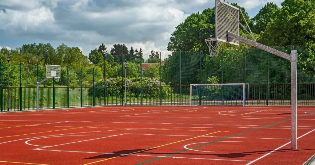 An outdoor red multi-purpose sports court featuring basketball hoops and a soccer goal surrounded by a high green fence.