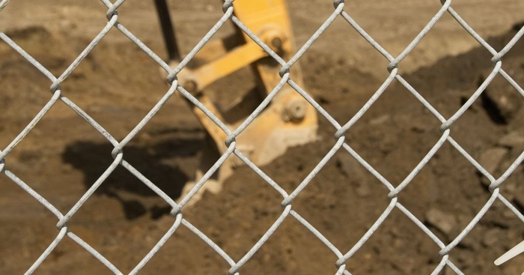 A close-up view through a chain-link fence showing a yellow excavator bucket digging into brown dirt.
