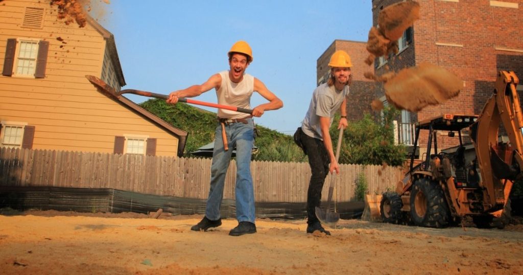 Two men in yellow hard hats posing enthusiastically with shovels at a construction site