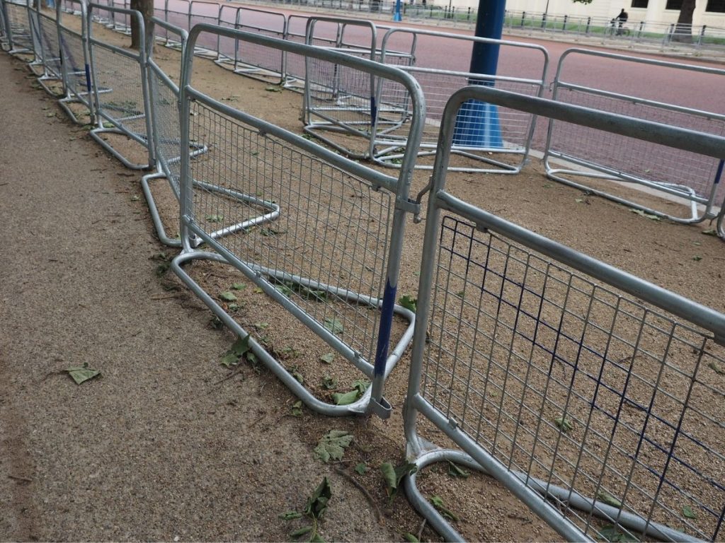 A line of interlocking grey metal crowd control barriers with wire mesh panels set up on a dirt and gravel path.
