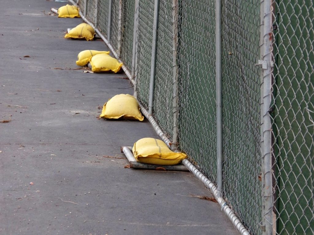 A line of yellow sandbags securing the base of a chain-link fence with green privacy screening on a grey asphalt surface.