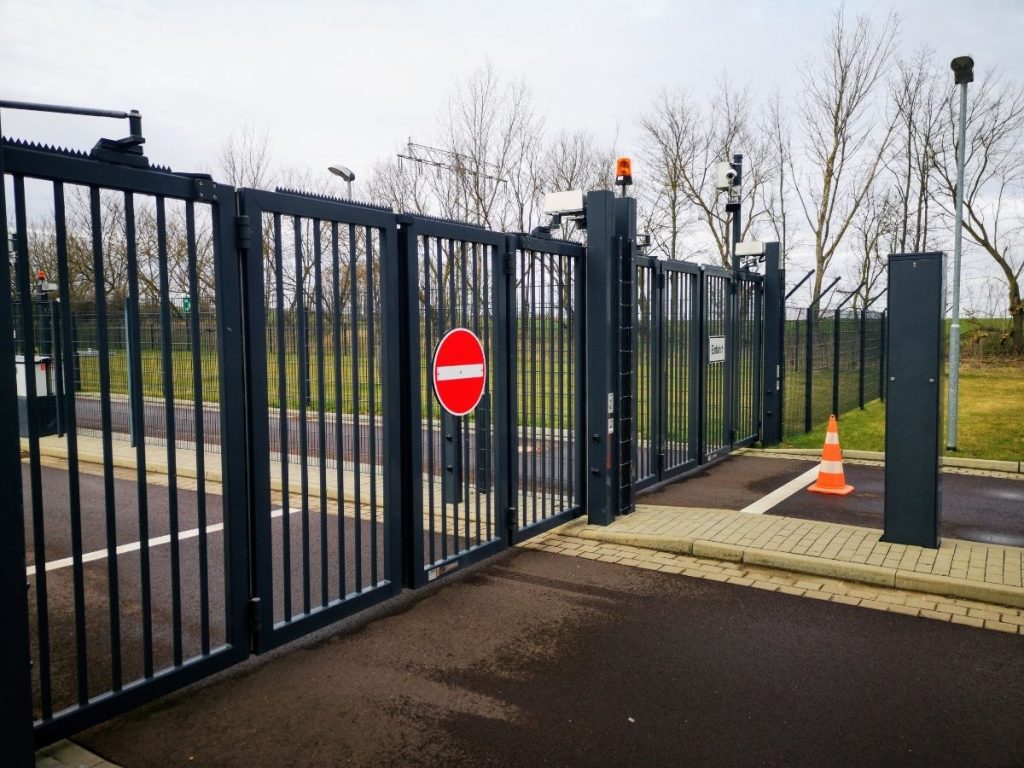 A high-security black metal folding gate at an entrance, featuring a "No Entry" sign, surveillance cameras, and an orange traffic cone.