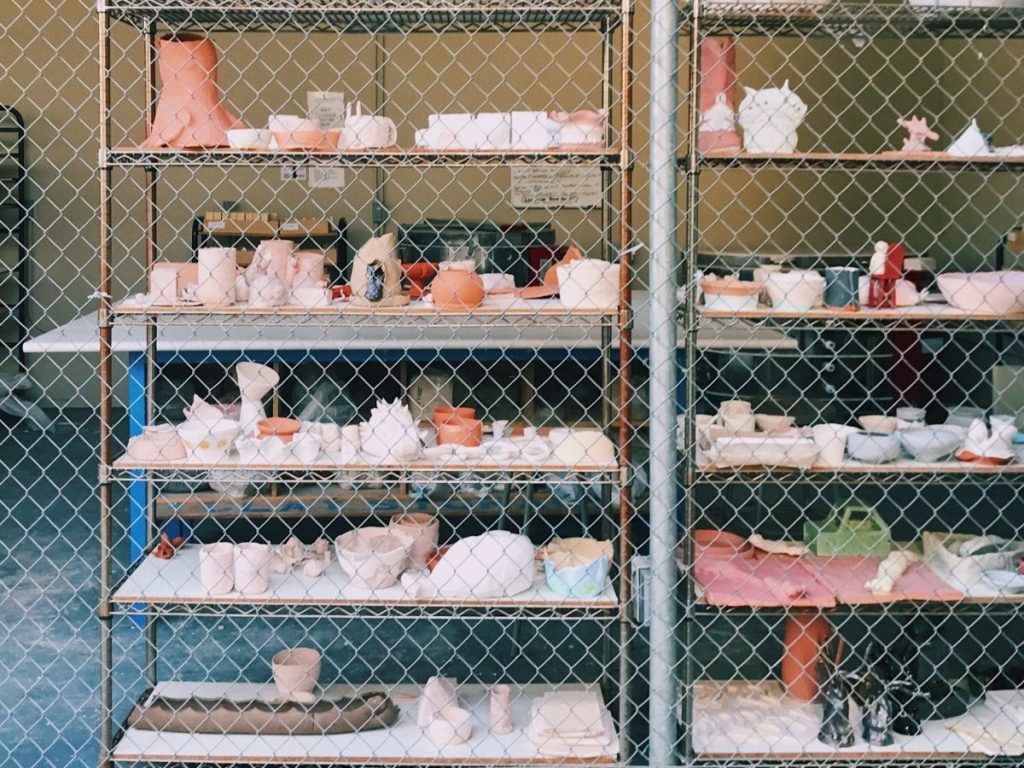 Various ceramic and clay pottery pieces displayed on industrial wire shelving units behind a chain-link security fence.