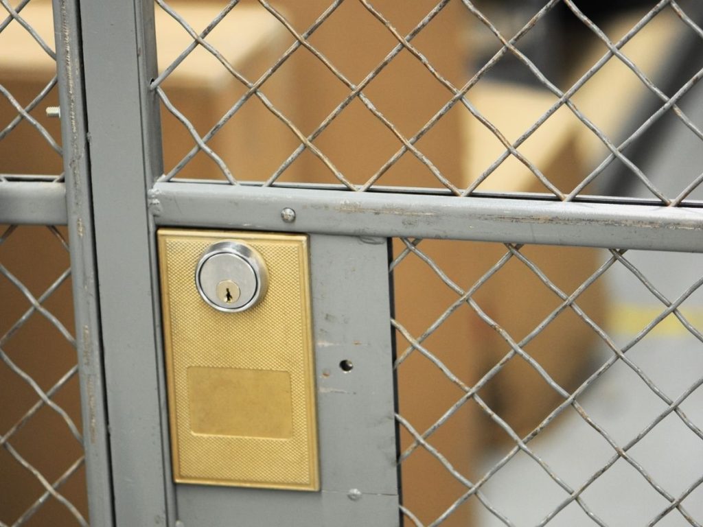 Close-up of a gold-coloured key lock cylinder and rectangular plate mounted on a grey metal wire mesh gate.