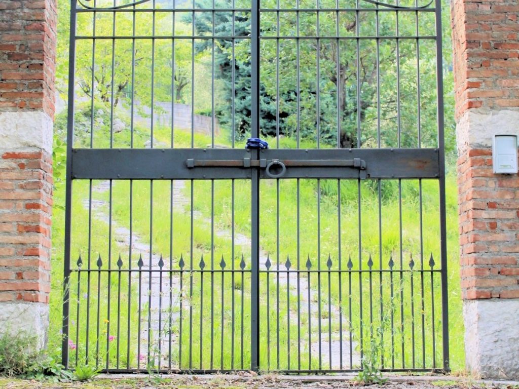 A black wrought-iron gate set between two brick pillars, revealing a grassy path and trees beyond.