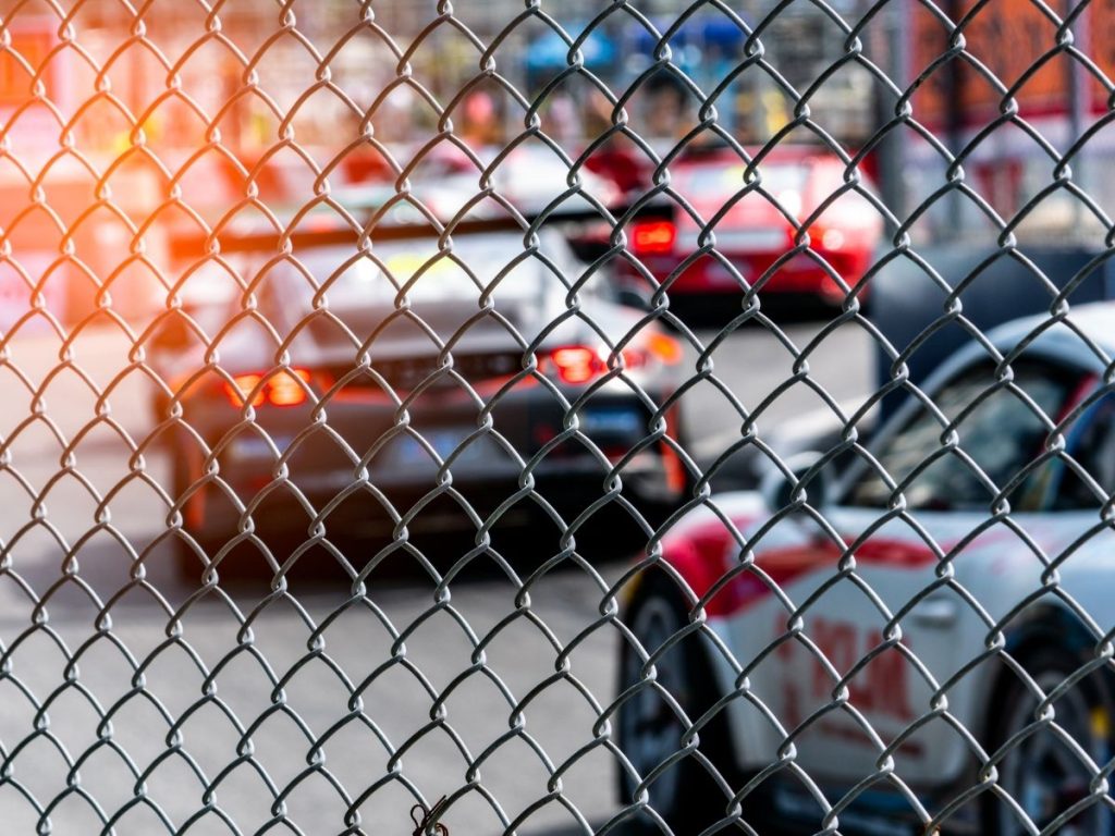 A close-up view looking through a chain-link fence at several race cars blurred in the background on a track.