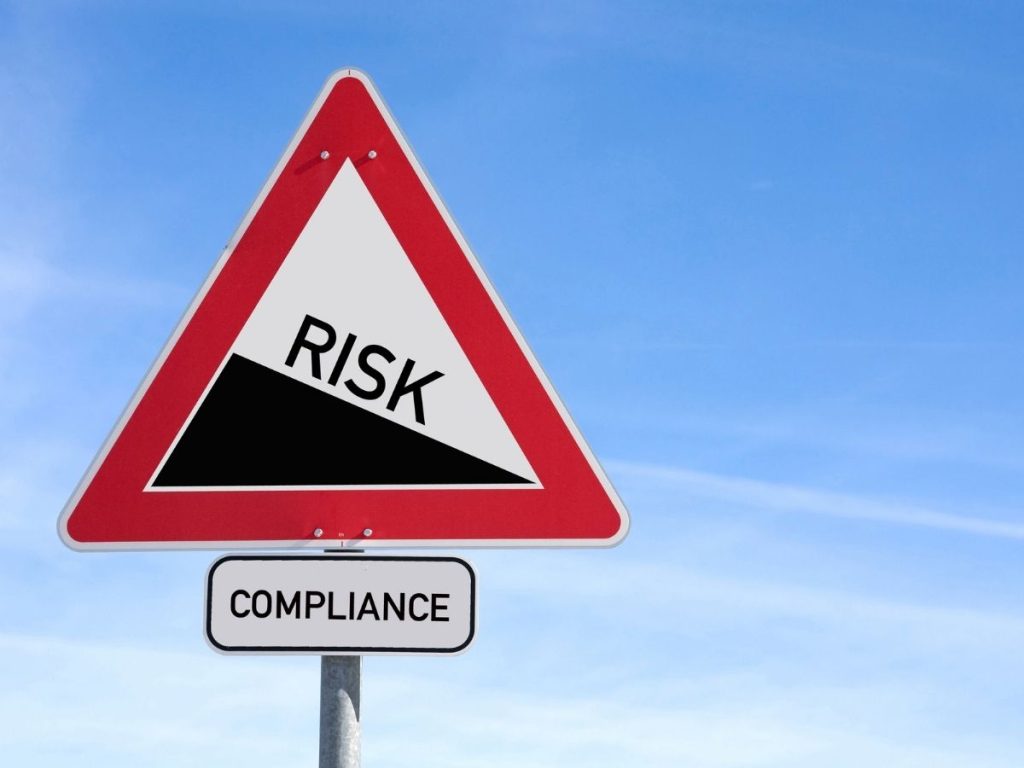 A triangular road sign with a red border containing the word "RISK" over a downward slope, with a smaller "COMPLIANCE" sign below it against a blue sky.