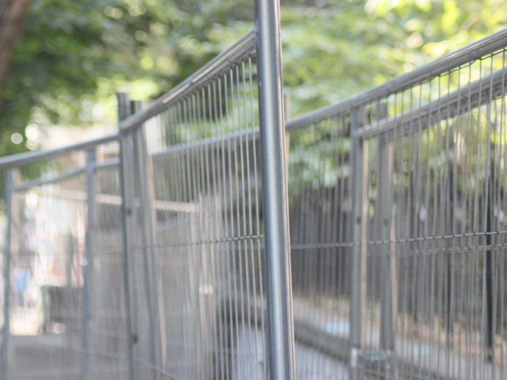 A close-up, slightly blurred shot of a curved metal security fence with vertical wire mesh, set against a background of soft green trees.