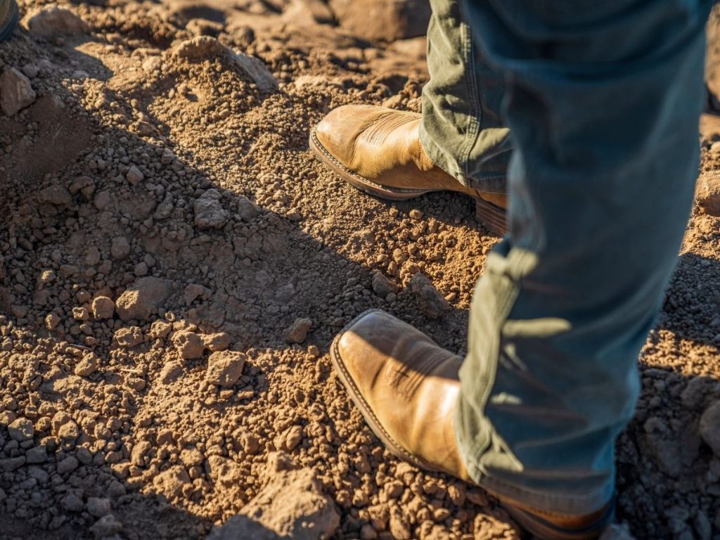 A person's feet wearing tan leather boots and green work pants, standing on dry, rocky soil.