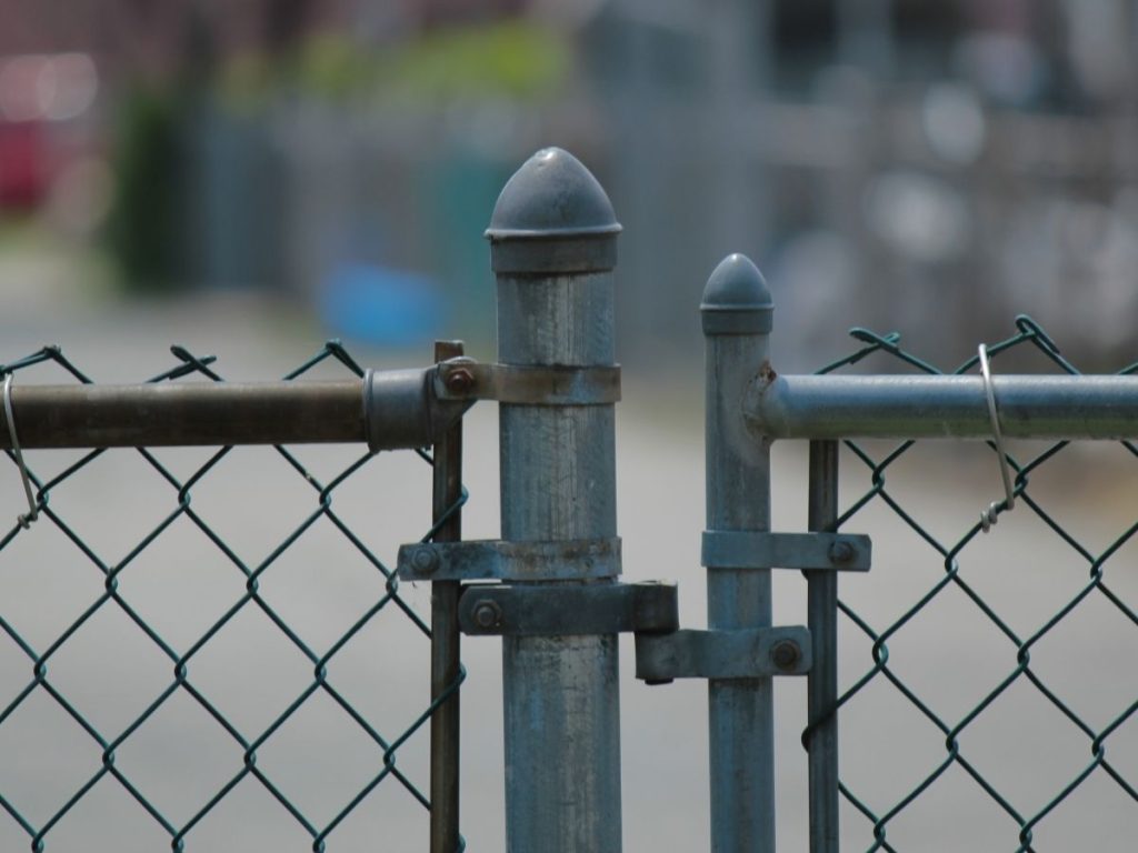 A close-up of a galvanised chain-link fence gate hinge and post with a blurred background.