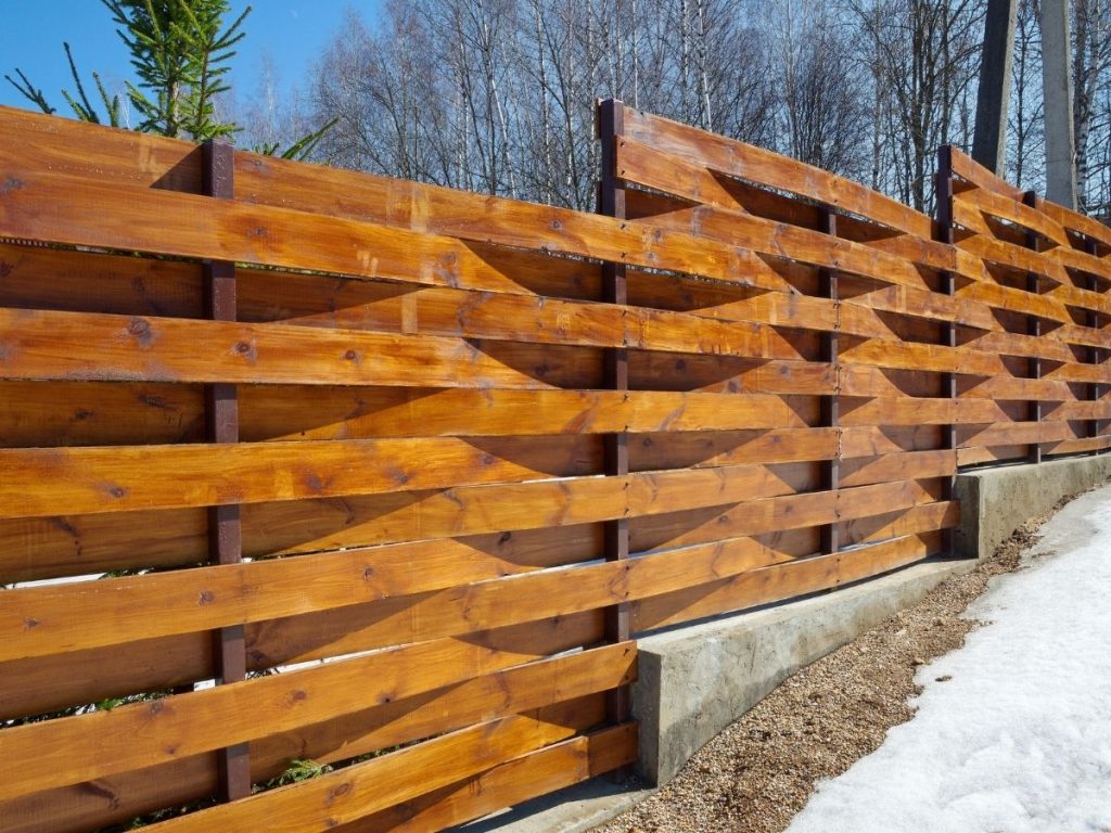 A horizontal, woven wooden fence made of warm-toned planks set on a concrete base, with a backdrop of bare winter trees and a clear blue sky.