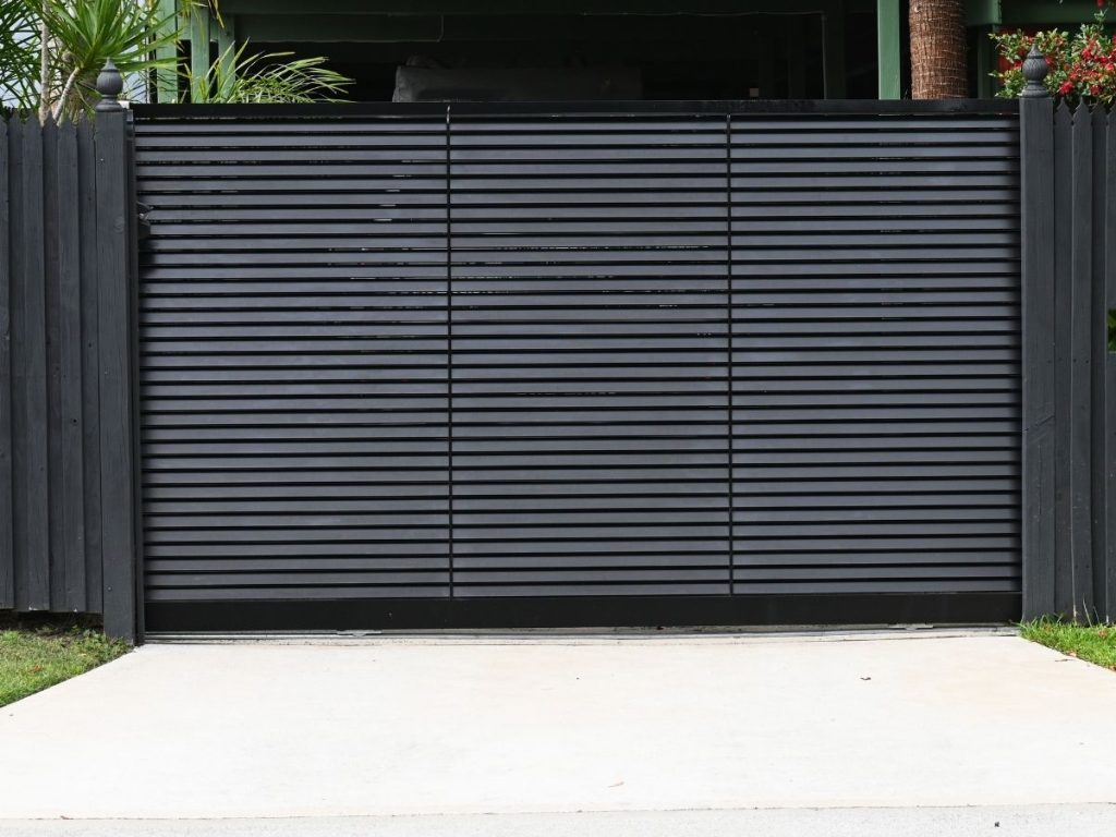 A modern black slatted driveway gate installed between two dark wooden fence posts on a concrete surface.